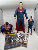 Three Superman action figures displayed: large 19" figure, medium 12" figure, and Superman bust bank with chain. Two comics in front.