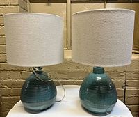Pair of two ceramic base table lamps with fabric drum shades on white round table against beige brick wall.