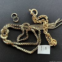 Gold tone chunky chain necklace, two long knotted box chain necklaces with tassels, and small taper hoop earrings on black background.