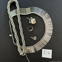 Five pieces of silver jewelry shown on a black background including a fringed metal pin choker, braided necklace, Maori fern pendant, pearl pendant, and sapphire zirconia ring.