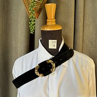 Front view of the black suede belt draped on a mannequin torso showing the large gold-tone decorative buckle and smooth suede texture.