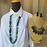 Long green malachite bead necklace displayed on a mannequin wearing a white shirt. A jewelry bust nearby holds a navy and gold teardrop choker necklace and matching earrings.