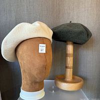 Cream and khaki green wool berets displayed on separate wooden hat stands against a fabric backdrop.