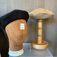Photo showing dark grey wool beret on tan mannequin head and camel wool beret on wooden hat stand.