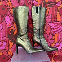 Side view of metallic gold-tone leather knee high boots with pointed toe and 3.5-inch heel against a floral background.