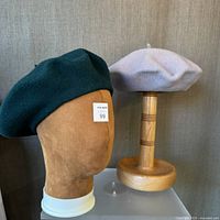 Two wool berets displayed on head forms: forest green Emanuel Geraldo beret and lavender beret side by side.