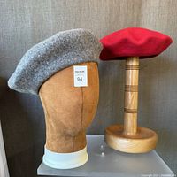 Two wool berets on head forms: grey on brown suede head form, red on wooden stand
