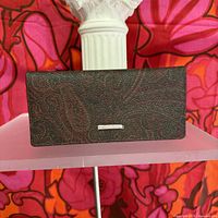 Full front view of a dark brown pebbled leather wallet with embossed paisley pattern, displayed on a pink shelf against a colorful floral background.
