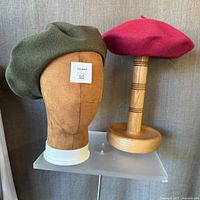 Both wool berets displayed on separate stands; khaki beret on a brown mannequin head, red beret on a wooden stand.