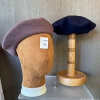 Two wool berets shown, mauve on a suede mannequin head and navy on a wooden stand, facing forward.