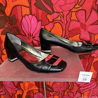Pair of black Marc Fisher heeled shoes with metallic band detail on heels and toes, displayed against a floral background.