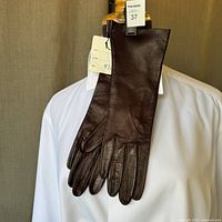 Full view of brown leather gloves hung on a hanger over a white shirt showing length and fingertips.
