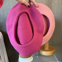 Two pink wool berets held with fingers, shown from the inside shape with visible hole openings, one darker pink and one lighter pink, displayed against a gray fabric background.