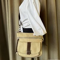 Full side view of the taupe pebbled leather Roots crossbody bag worn on mannequin, showcasing dark brown leather strap and flap pocket.