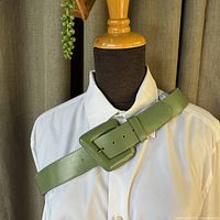 Full view of the sage green leather belt displayed on a mannequin wearing a white shirt, showing the angle and placement of the buckle.
