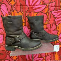 Side profile of one brown leather boot on clear stand showing the strap with buckle and heel