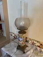 Full view of antique electric parlor lamp showing clear etched glass chimney, painted middle glass globe with winter cabin and autumn leaves, brass claw-foot base, and electric cord.