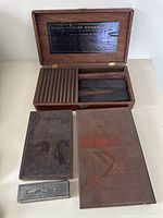 Photo showing opened wooden box with compartments and two advertising etched plates partially visible along with a metal tool on the side.