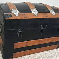 Side and front view of the vintage blanket chest showing black exterior with wood strapping and metal fasteners on domed top and body, lock clasps visible, handles missing.