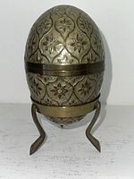 Front view of brass egg-shaped box on curved stand, showing embossed floral detailing.