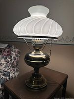 Front angled view of antique brass hurricane lamp with white swirl milk glass shade and clear glass chimney on wooden table next to floral-patterned sofa.