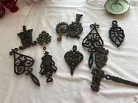 Top-down view of all vintage cast iron trivets and one wall hanger hook arranged on a white cloth showing detailed shapes and openwork designs.