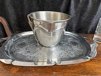 Front view of polished stainless steel ice bucket sitting on ornate engraved metal tray