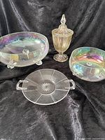 Full view of all four iridescent glass items on black fabric background showing colors and shapes of bowls, plate, and candy dish with lid.