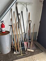 Photo of collection of long handled lawn and garden tools leaning against wall including rakes, shovels, pitchfork, drywall tool, brush, and child’s snow shovel.