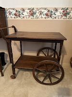 Full front view of antique oak tea cart showing top surface, large spoked wheels on one side, small caster wheels on opposite side, and turned legs.