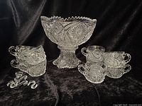 Pedestal punch bowl, 10 matching cups and metal hooks arranged on black cloth, showing clear ornate cut glass design and zig zag edge of the bowl.