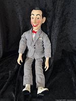 Full length photo of the Pee Wee Herman doll sitting, showing grey checkered suit and white shoes on black background