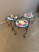 Front view of three metal plant stands with different fruit and floral ceramic tile tops on a carpeted floor against a wall.