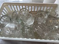 Top view of a white plastic basket full of various clear glass salt cellars showing diversity of shapes and designs