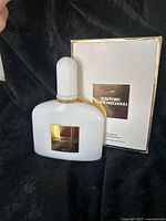 White ribbed perfume bottle with gold label and original box in background on black fabric.