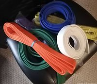 Six martial arts belts in blue, green, orange, purple, white, and yellow colors arranged together, some wrapped in plastic.