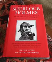 Front cover of The Complete Sherlock Holmes hardback book showing title, author, and illustration of Sherlock Holmes with pipe and cap on red background.