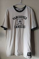 Front view of white cotton t-shirt with black collar and sleeve edges. Shows 'Vancouver Canucks' text and logo.