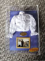 Front view of the clear acrylic display case showing the white puffy shirt replica mounted on a purple background with Seinfeld branding, a gold plaque, and a photo from the episode.