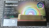 Box front showing the Brookstone Rainbow LED Ambient Light with a depiction of the product displaying multicolor, dancing LEDs in a rainbow shape on its wood-look base. Text mentions sound reactive and color changing features.
