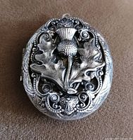Front of the oval locket pendant showing raised thistle flower and leaves in relief against ornately patterned background.