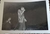 Close-up black and white image of Elvis Presley performing live in Toronto 1957 wearing his gold lame jacket, holding a microphone, side view on stage with crowd visible.