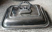 Rectangular silver-plated serving tray with ornate lid and decorative handle shown closed on carpeted floor