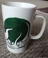 Close-up photo of white ceramic mug showing artistic image of a white cat playing with a large green ball of yarn on the side of the mug.