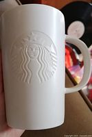 Front side of the tall white ceramic Starbucks mug showing embossed siren logo with a matte finish and handle.