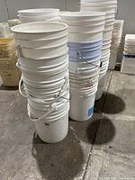 Side view of stacked white 5-gallon buckets showing some with metal handles and some without on concrete floor.