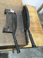 Side view of two heavy metal floral stem cutters mounted on a wooden table surface, showing black padded handles and robust cutting bases.