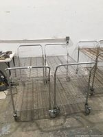 Front view of two metal wire utility carts side by side showing their handles and two-tiered shelving.