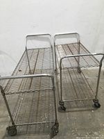 Photo showing two metal wire utility carts side by side with handles and locking wheels, exhibiting rust spots.