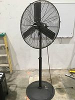 Full view of the 30" black metal pedestal fan against a warehouse wall with a utility ladder nearby.